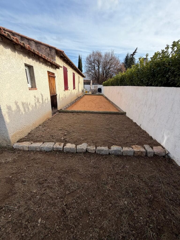 Terrain de pétanque aménagé avec surface stabilisée et bordures en bois le long d’une maison.
