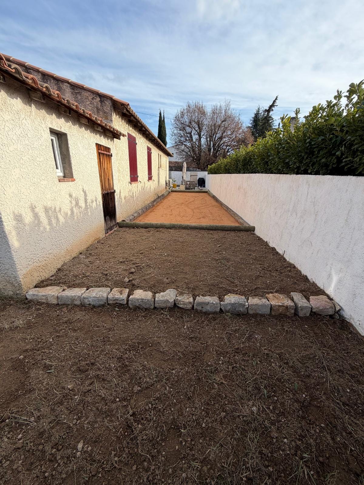 Terrain de pétanque aménagé avec surface stabilisée et bordures en bois le long d’une maison.