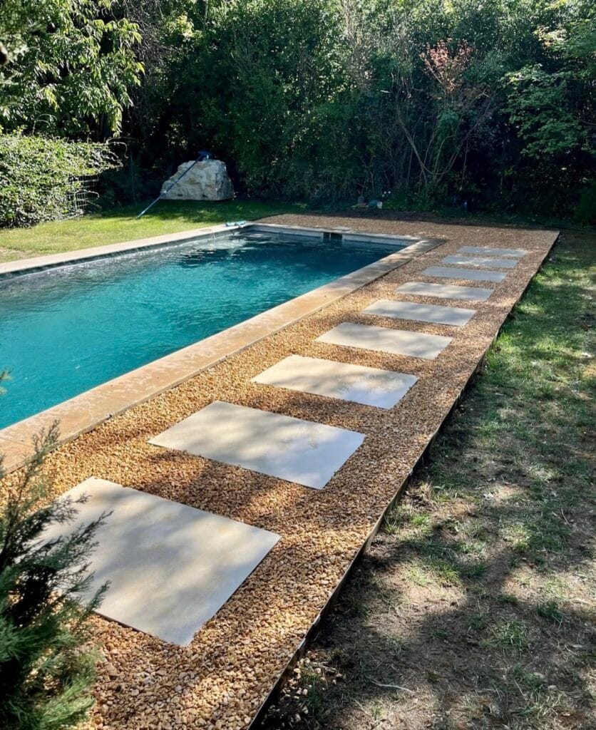 Chemin de dalles entourant une piscine moderne avec margelles claires et gravier, aménagé dans un jardin verdoyant.