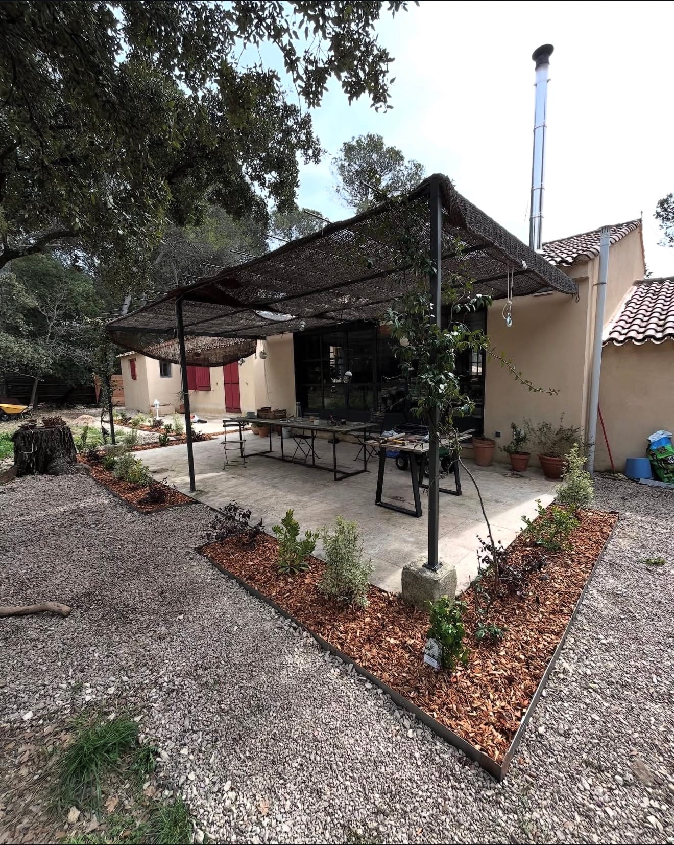 Terrasse en dalles claires avec allée pavée, zones de plantations et bordure de jardin devant une maison, dans un aménagement paysager extérieur.