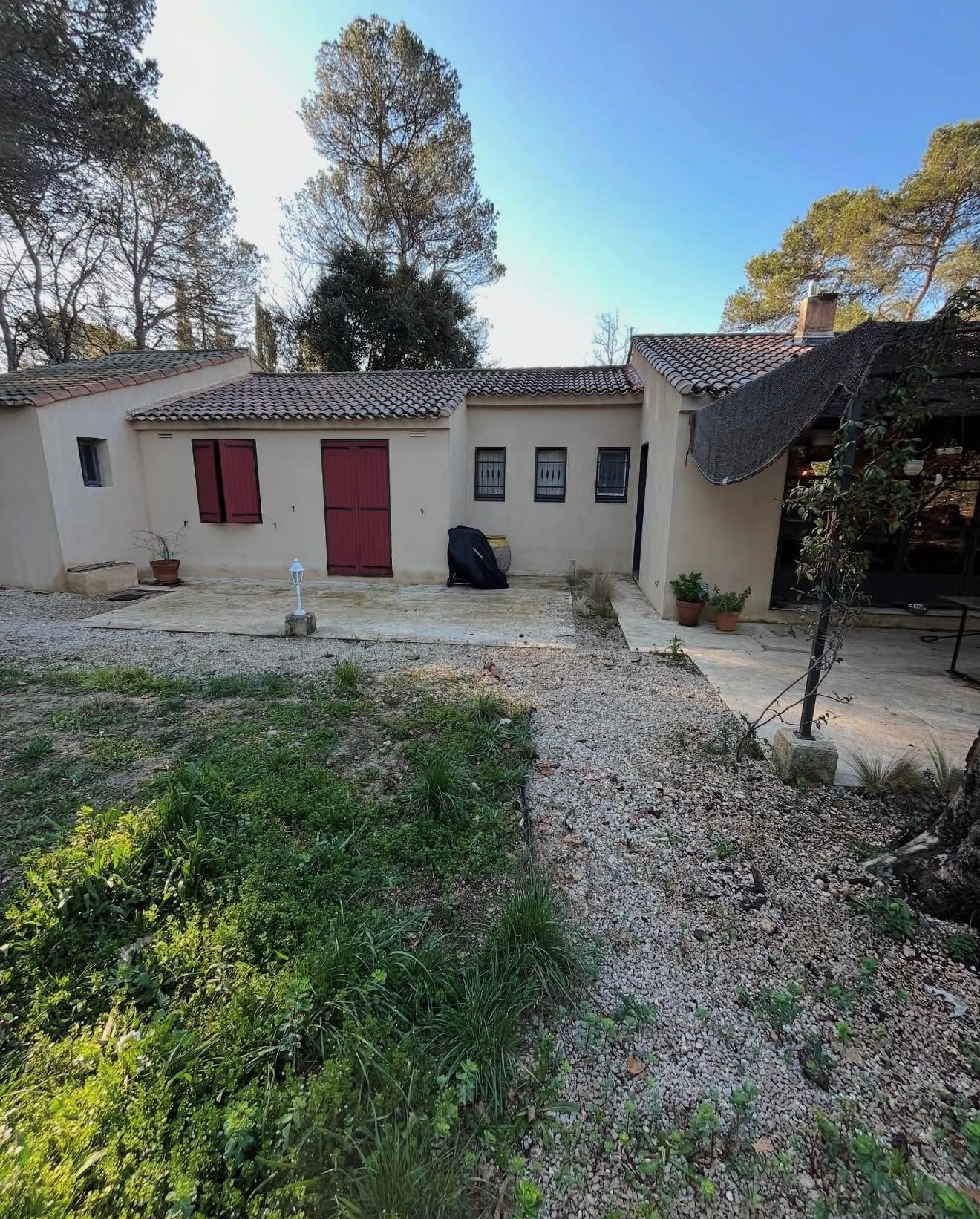Façade d’une maison en pierre claire avec zone de terrasse, jardin et allée en gravier, entourée de pins et végétation.