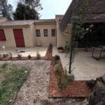 Aménagement paysager de terrasse et jardin devant une maison beige avec allées pavées et massifs plantés bordés de paillis.
