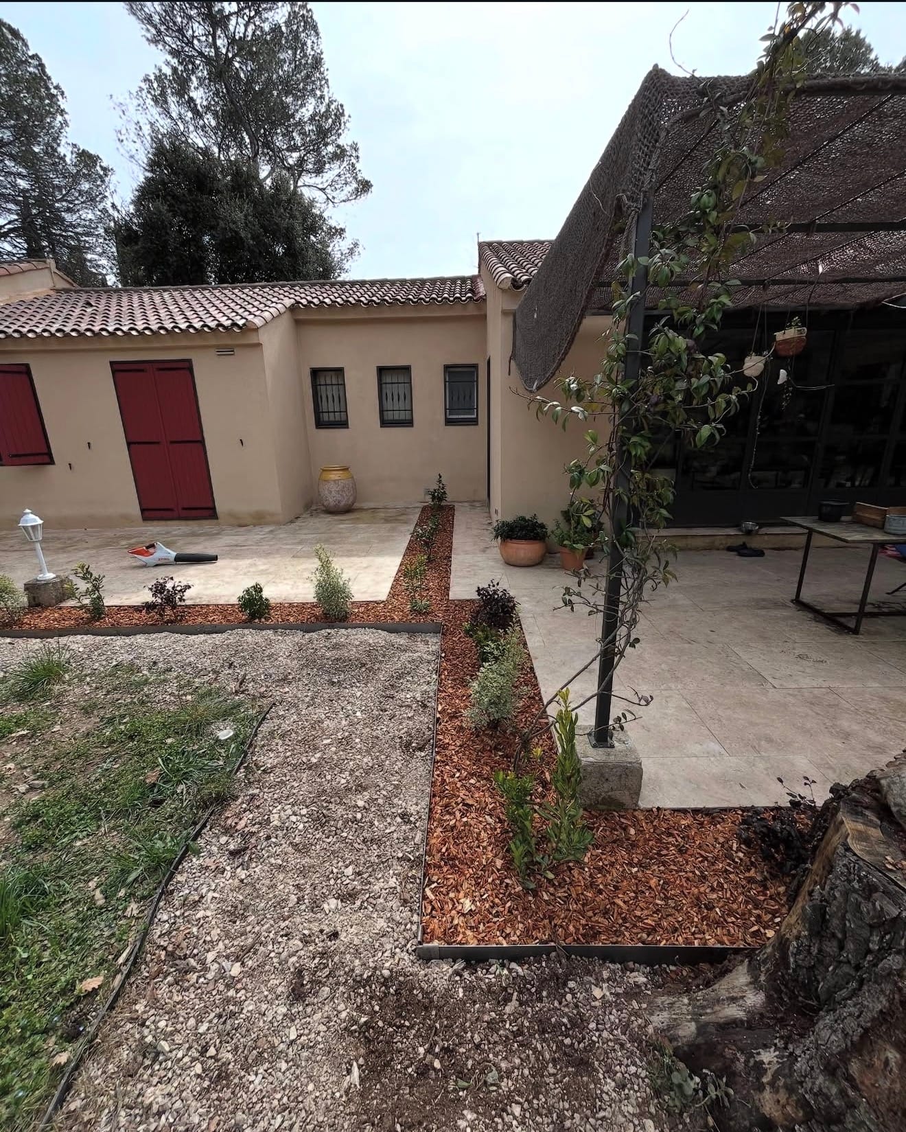 Aménagement paysager de terrasse et jardin devant une maison beige avec allées pavées et massifs plantés bordés de paillis.